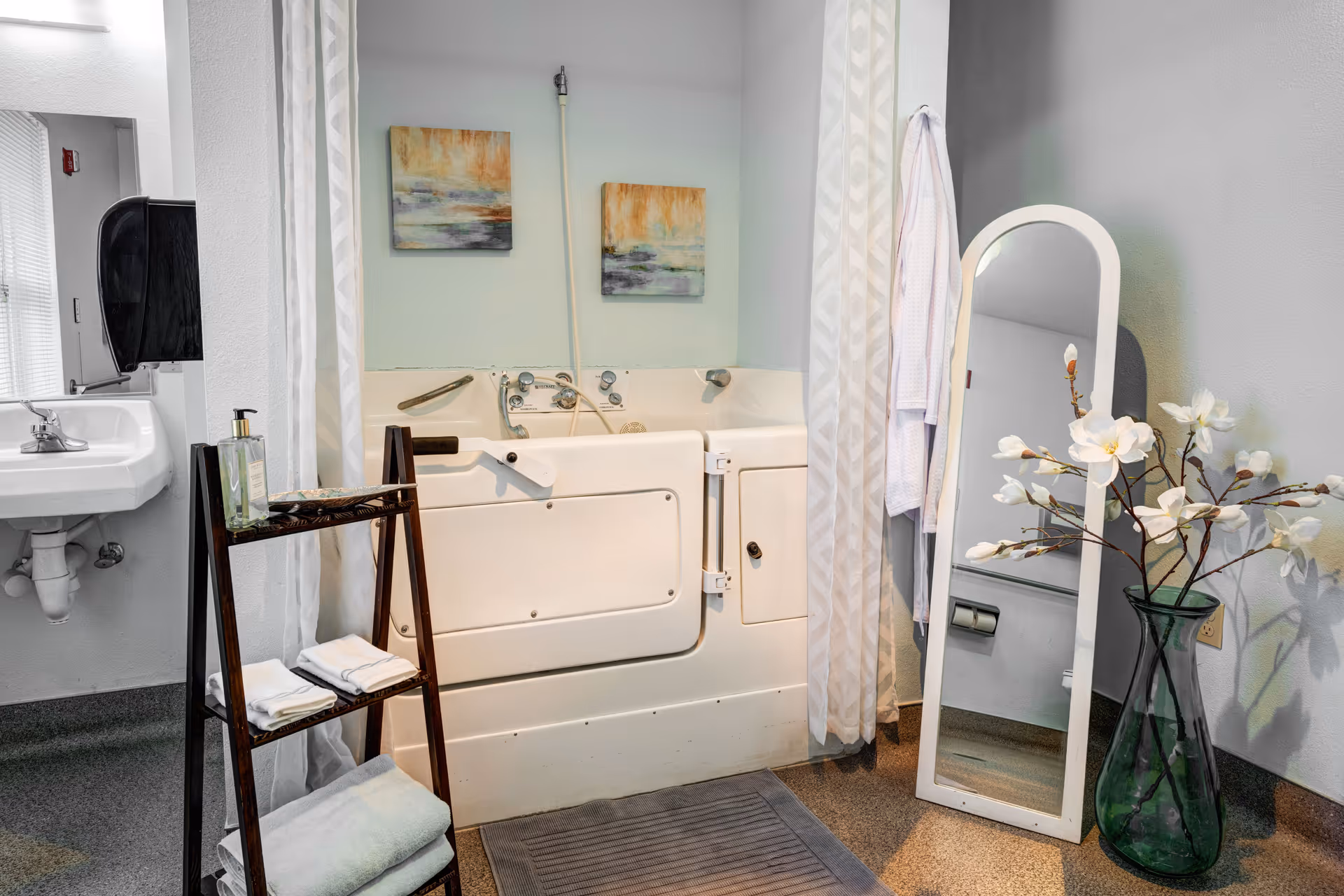 Walk-in bathtub in an assisted-living bathroom with a small sink, towel ladder, freestanding mirror, and vase of flowers.