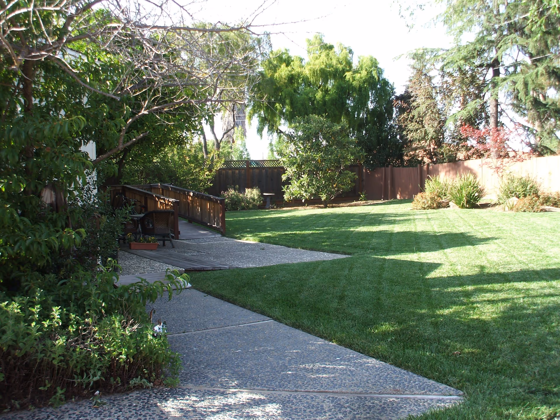 A well-maintained outdoor garden area with a paved walkway, green grass, various trees and shrubs, and a wooden fence in the background. There is a wooden ramp with railings and some outdoor chairs near the left side.