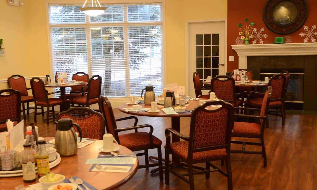 Dining room with several round wooden tables and red cushioned chairs arranged around them. Tables are set with coffee pots, cups, menus, and condiments. Large windows with white blinds allow natural light to fill the room. A fireplace with a mantel decorated with a clock and seasonal decorations is visible on the right wall.