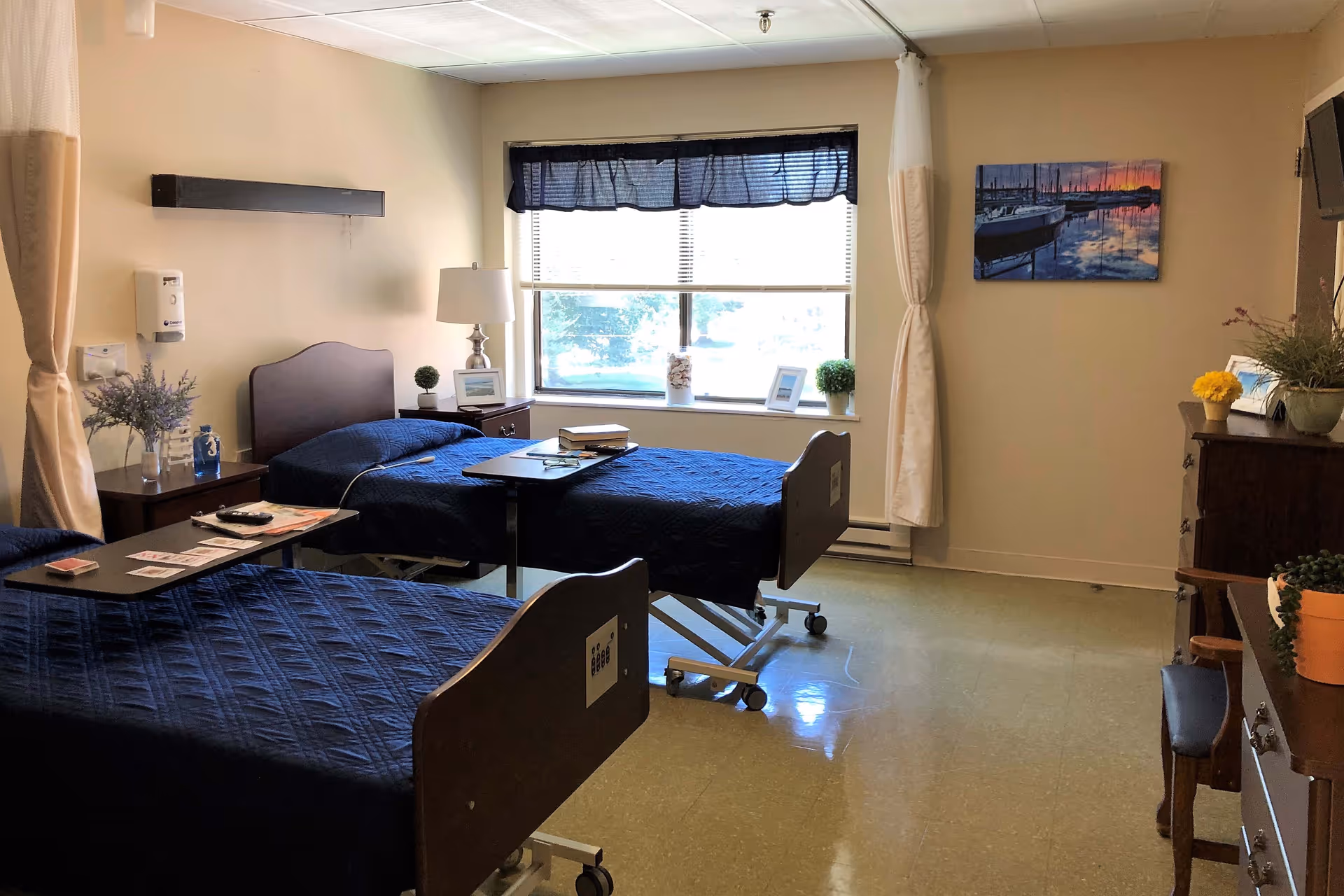 A shared patient room in a healthcare facility with two hospital beds covered in dark blue quilts. Each bed has an overbed table with items such as books, glasses, and playing cards. The room has a large window with a dark blue valance, a nightstand with a lamp and small plants, a dresser with flowers and framed pictures, and a wall-mounted TV. The walls are beige, and there is a curtain that can be drawn for privacy between the beds.