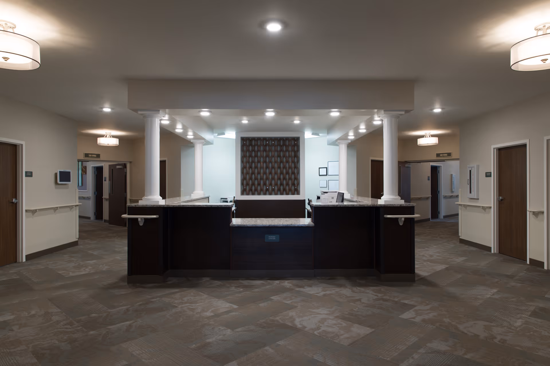 A spacious reception or nurse station area in a senior living facility with a dark wood counter, white columns, and neutral-colored walls and carpet. The area is well-lit with ceiling lights and has multiple doorways leading to other rooms or hallways.