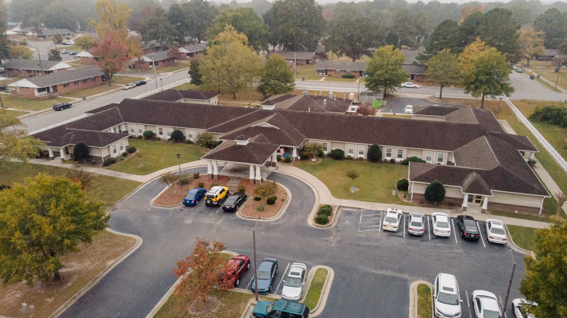 Aerial view of a single-story senior living facility with a covered entrance, circular driveway and parked cars surrounded by trees and nearby houses.