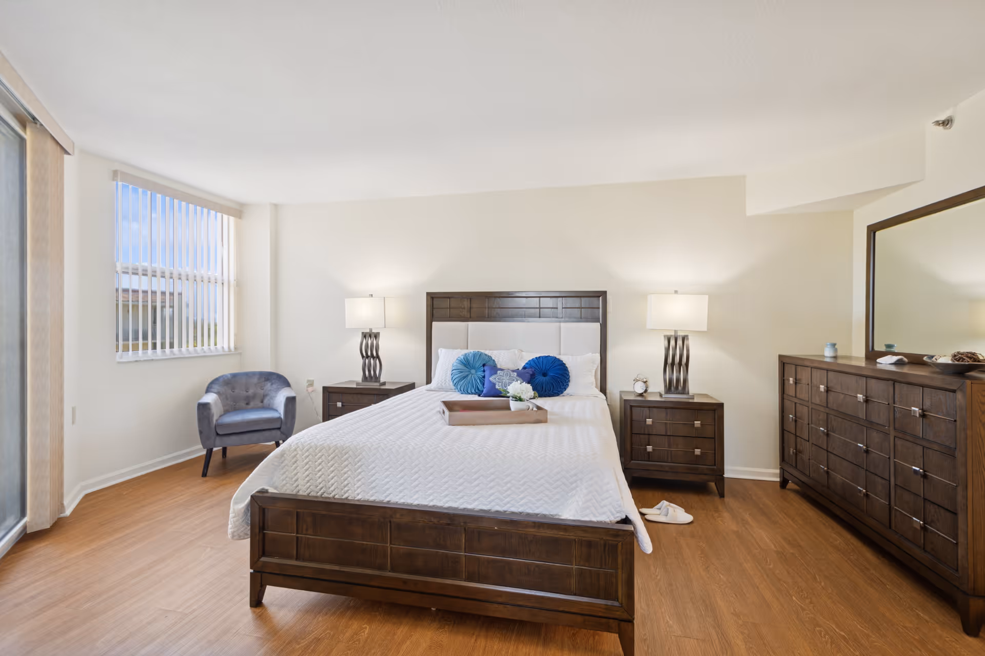 A bright bedroom with a wooden bed frame and white bedding, decorated with blue and white pillows. There are two matching wooden nightstands with lamps on each side of the bed. A wooden dresser with a large mirror is on the right side of the room. A gray upholstered chair is positioned near a window with vertical blinds. The floor is a light wood finish.