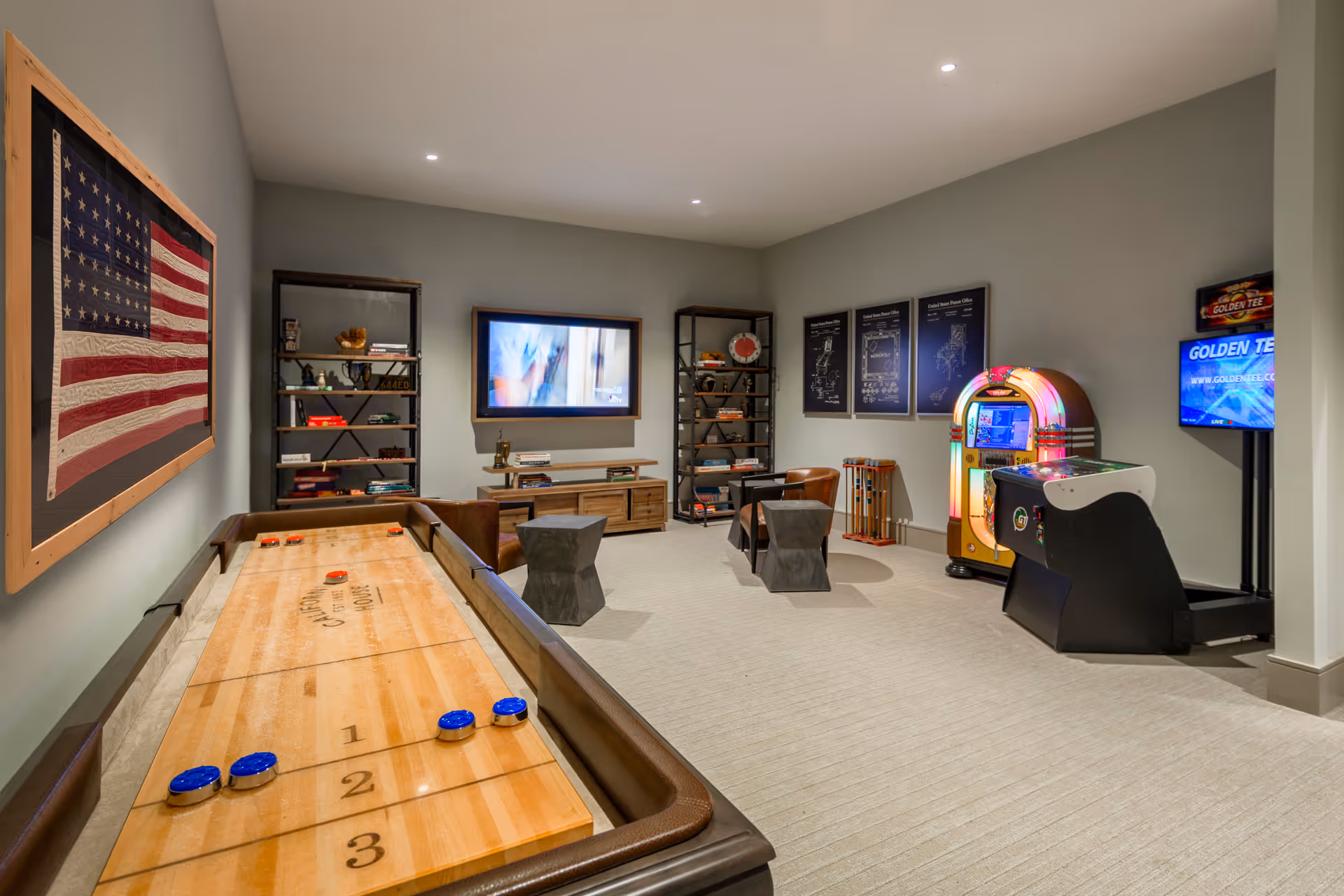 A recreational room with a shuffleboard table in the foreground, an American flag artwork on the left wall, two black shelving units with decorative items, a wall-mounted TV, two modern chairs with small side tables, a vintage jukebox, and an arcade game machine on the right side.