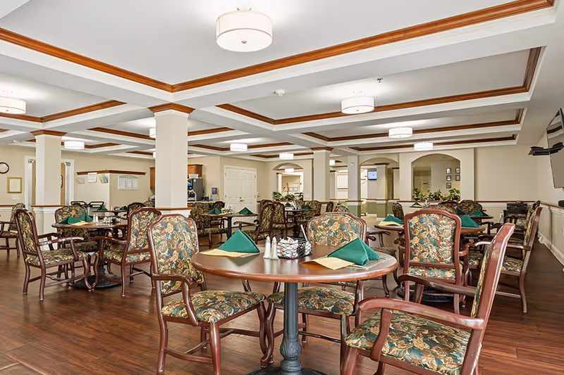 A spacious dining room with multiple round wooden tables and floral upholstered chairs. Each table is set with green folded napkins, menus, and condiment holders. The room features a coffered ceiling with recessed lighting, cream-colored walls, and wooden flooring. There are columns and archways in the background, along with some plants and a wall-mounted television.