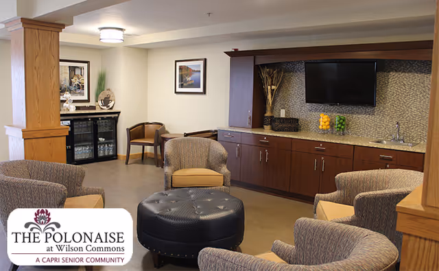 A cozy senior living community lounge area with four upholstered armchairs arranged around a large round black ottoman. The room features wooden cabinetry with a mounted flat-screen TV, a small sink, and decorative items including jars of lemons and limes. There is a small table with two chairs and framed artwork on the walls. The space has warm lighting and a welcoming atmosphere.