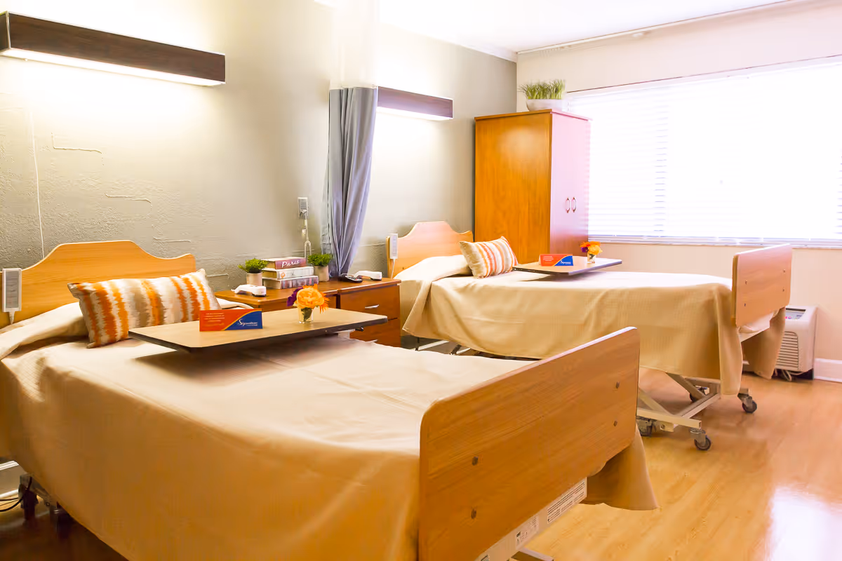 A bright shared patient room with two neatly made hospital-style beds, overbed trays, bedside tables, and a window with blinds.