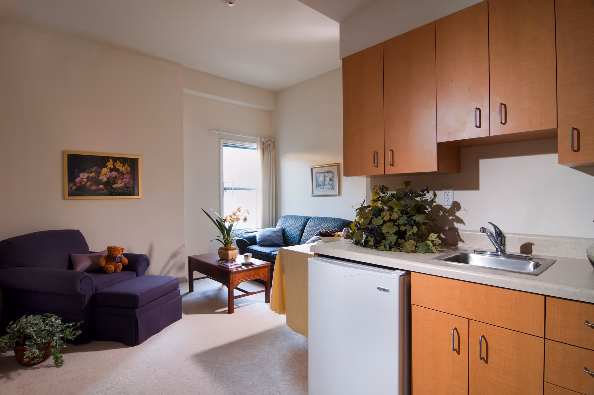 A cozy living area with a purple armchair and ottoman, a small wooden coffee table with a potted plant and books, a blue couch near a window with curtains, and a kitchenette with wooden cabinets, a sink, and a small refrigerator. There are framed pictures on the walls and some greenery on the counter.