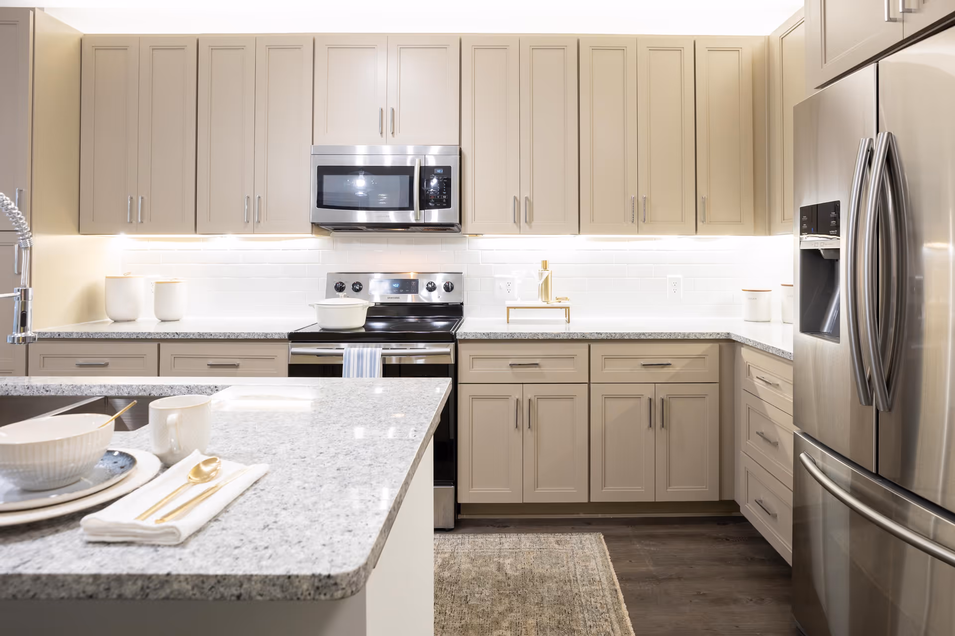 Modern kitchen with beige cabinets, stainless steel refrigerator, microwave, and stove. Granite countertops with a bowl, cup, and gold utensils on a white napkin are visible on the kitchen island. The backsplash is white subway tile with under-cabinet lighting.