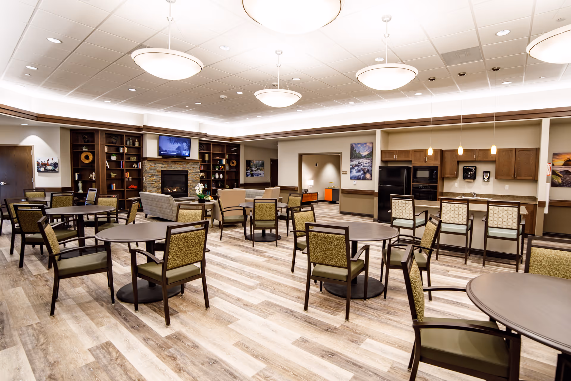 A spacious and well-lit common area in a senior living facility featuring multiple round tables with chairs, a seating area with armchairs and a sofa near a stone fireplace with a mounted TV above it, built-in wooden shelves with decorative items, and a kitchen area with a refrigerator, oven, and bar stools. The room has wood-patterned flooring and modern ceiling lights.