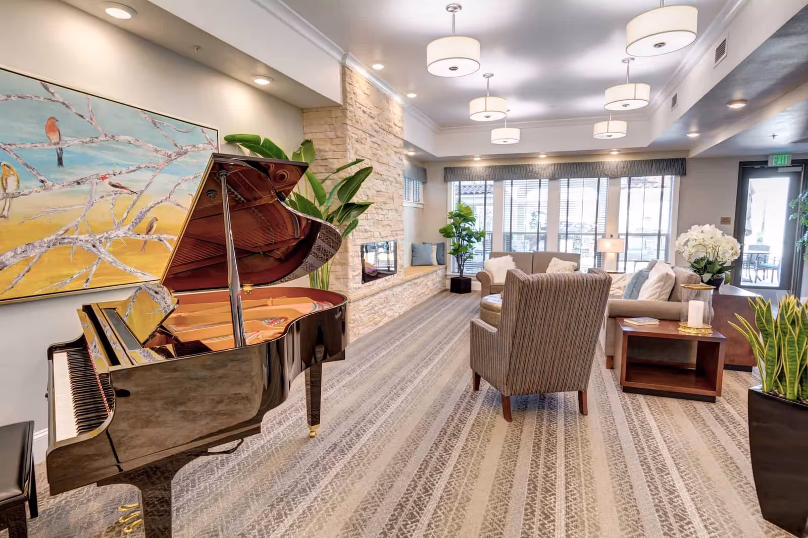 A spacious and well-lit living room area in Rose Arbor Village featuring a grand piano on the left, a large painting of birds on a branch above the piano, a stone fireplace along the back wall, comfortable seating including sofas and an armchair, several potted plants, and large windows with blinds letting in natural light.