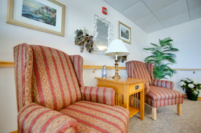 Two striped upholstered armchairs with red and beige patterns are positioned on either side of a wooden side table with a lamp and a small decorative item. The wall behind features two framed landscape paintings, a decorative mirror, and wall-mounted floral arrangements. A tall green potted plant and a bouquet of white flowers are placed in the corner of the room.