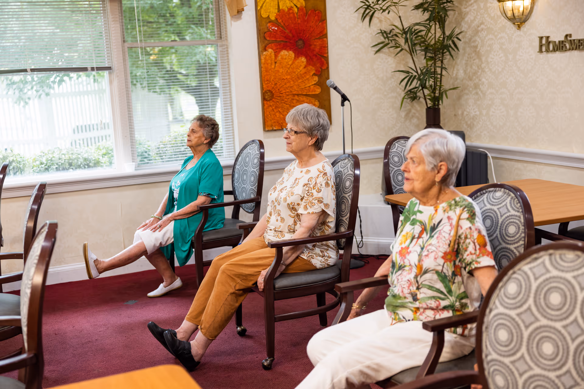 Three elderly women seated in chairs in a room with large windows, performing leg exercises by extending one leg forward. The room has patterned chairs, a red carpet, a plant, and a wall decoration with large orange and red flowers.