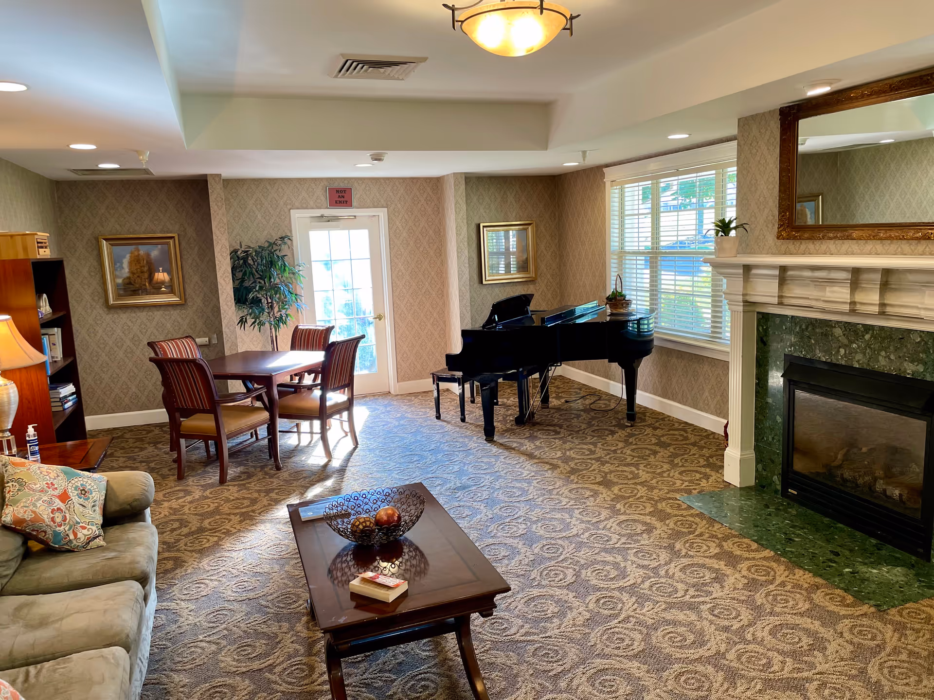 A cozy living room area with a beige patterned carpet, a green sofa with decorative pillows, a wooden coffee table with a decorative bowl and a book, a grand piano near a window with white blinds, a fireplace with a green marble surround and a large ornate mirror above it, a wooden table with four chairs near a glass door, and a bookshelf with a lamp and books.