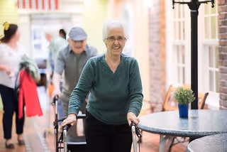 An elderly woman using a walker is smiling and walking indoors in a senior living facility. In the background, other elderly individuals and a caregiver are visible, along with a table and chairs in a hallway with warm lighting.