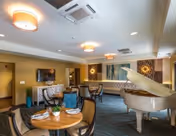 A spacious and well-lit living room area in a senior living facility featuring a white grand piano, round tables set with plates and napkins, comfortable chairs, a wall-mounted flat-screen TV, and decorative wall art. The room has a modern ceiling with recessed lighting and air vents.