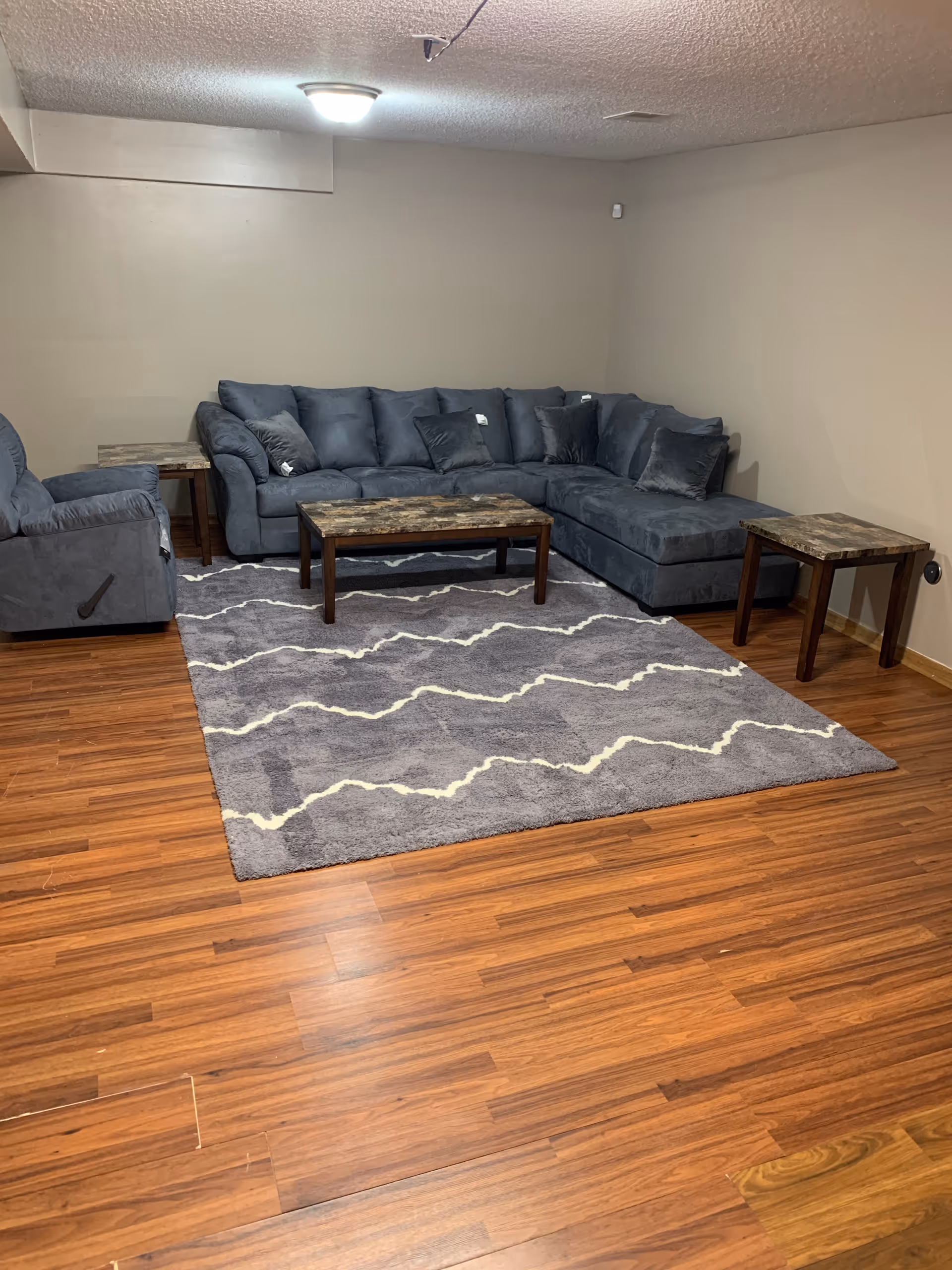 A living room with a large gray sectional sofa, a matching armchair, a patterned gray area rug, and three wooden tables with marble-like tops. The room has wood flooring and beige walls with a ceiling light fixture.