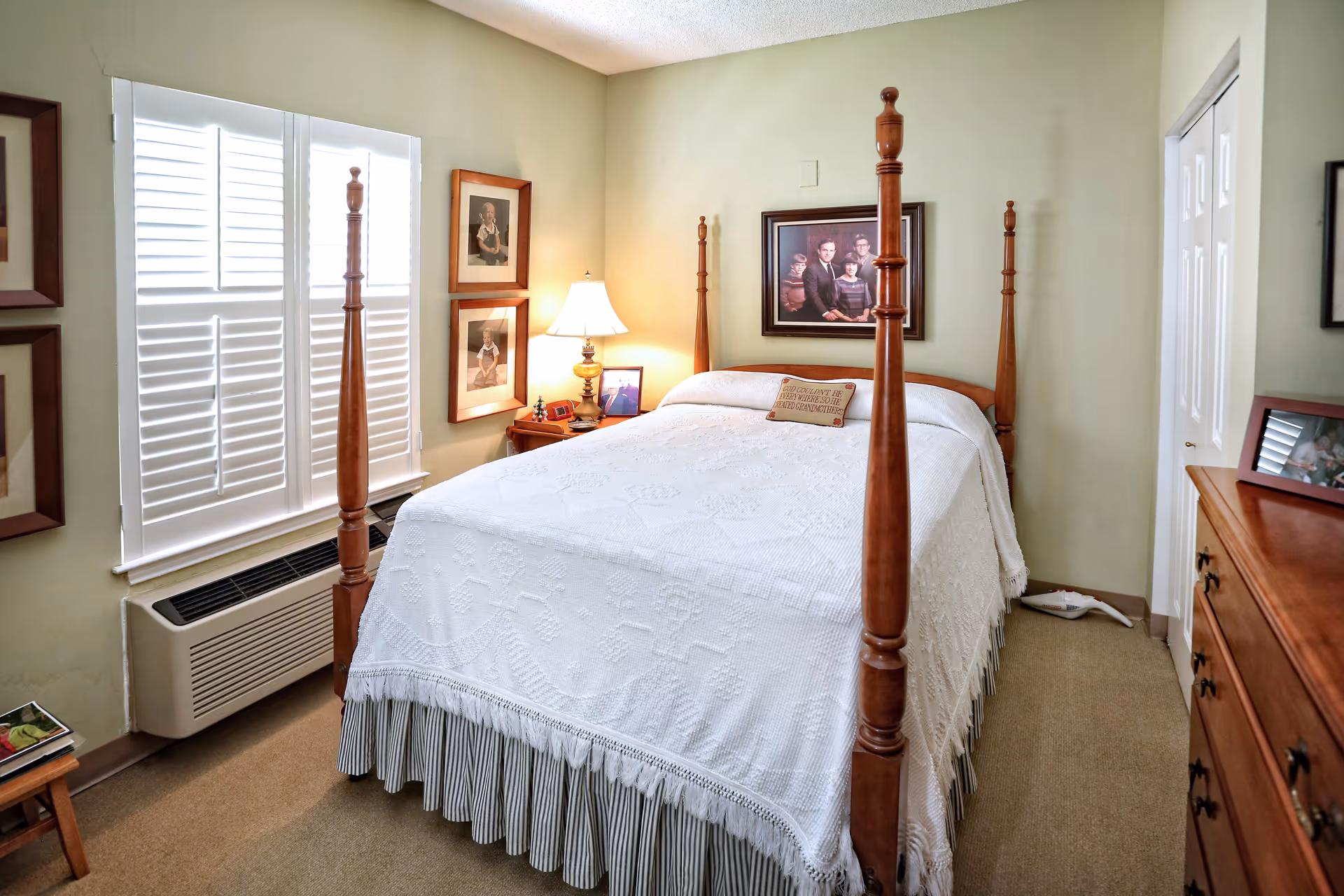 Traditional bedroom featuring a wooden four-poster bed with a white coverlet, nightstand and lamp, shuttered window, and dresser.