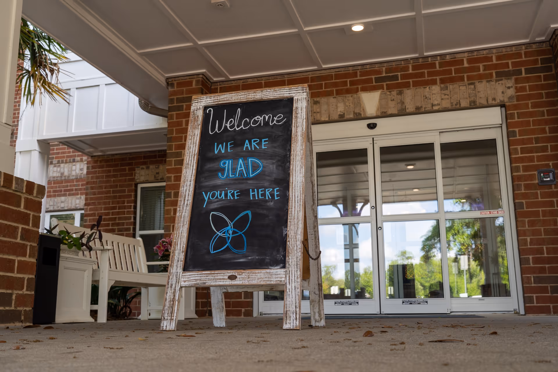 Entrance of a brick building with a covered porch. A wooden framed chalkboard sign stands on the ground with the message 'Welcome WE ARE GLAD YOU'RE HERE' written in white and blue chalk. There is a white bench and some plants to the left of the entrance. The glass doors reflect trees and the sky outside.