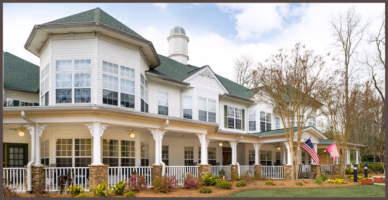 Exterior view of a large, two-story senior living facility building with white siding, green roof, multiple windows, a wraparound porch with white railings and columns, and two flags displayed near the entrance. The surrounding area includes landscaped plants and trees under a partly cloudy sky.