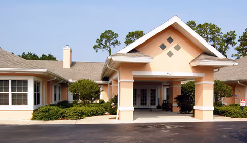 Front entrance of a low-rise peach-colored building with a covered porte-cochere, columns, shrubs, and a driveway.