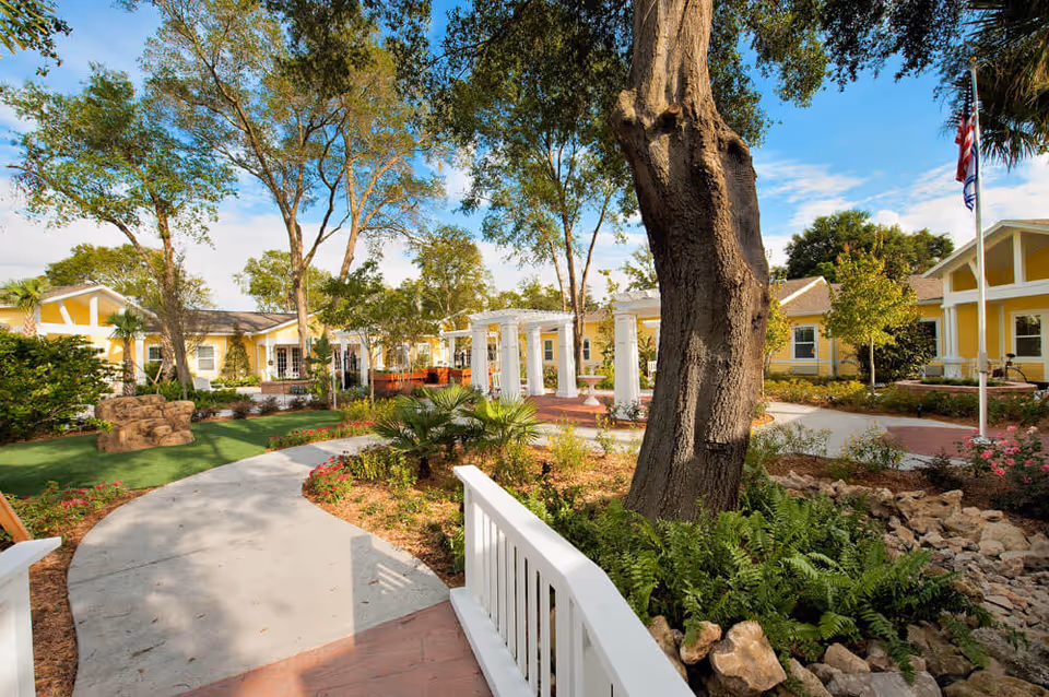A sunny outdoor garden area with a winding concrete pathway, large trees, green shrubs, and colorful flowers. Yellow buildings with white trim surround the garden, and there is a white pergola structure in the center. An American flag is visible on a flagpole to the right.
