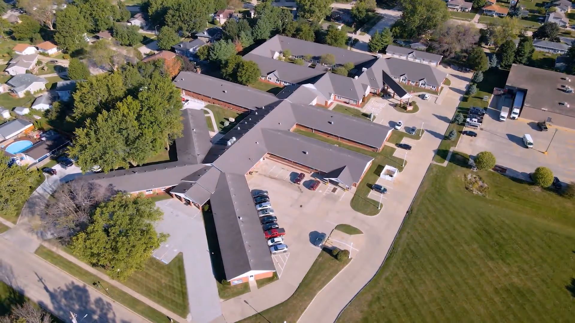 Aerial view of Regency Assisted Living facility showing multiple connected single-story buildings with gray roofs, surrounded by trees, parking lots with cars, and nearby residential houses.