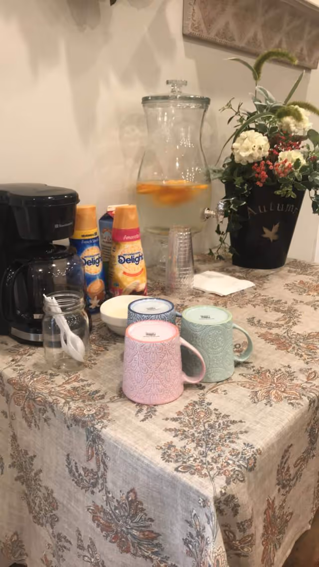 Table set up as a coffee station with a coffee maker, creamer bottles, decorative mugs, a glass water dispenser with fruit, and a floral arrangement.