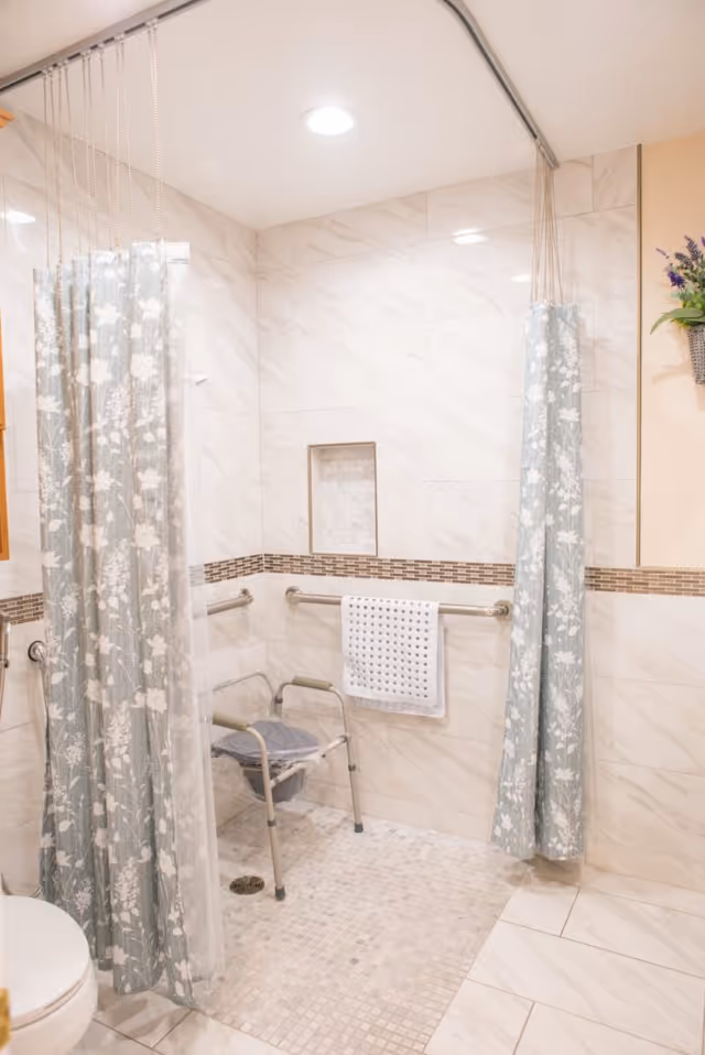 Accessible tiled shower with a folding shower chair, grab bars, a towel on a rail, and floral shower curtains.