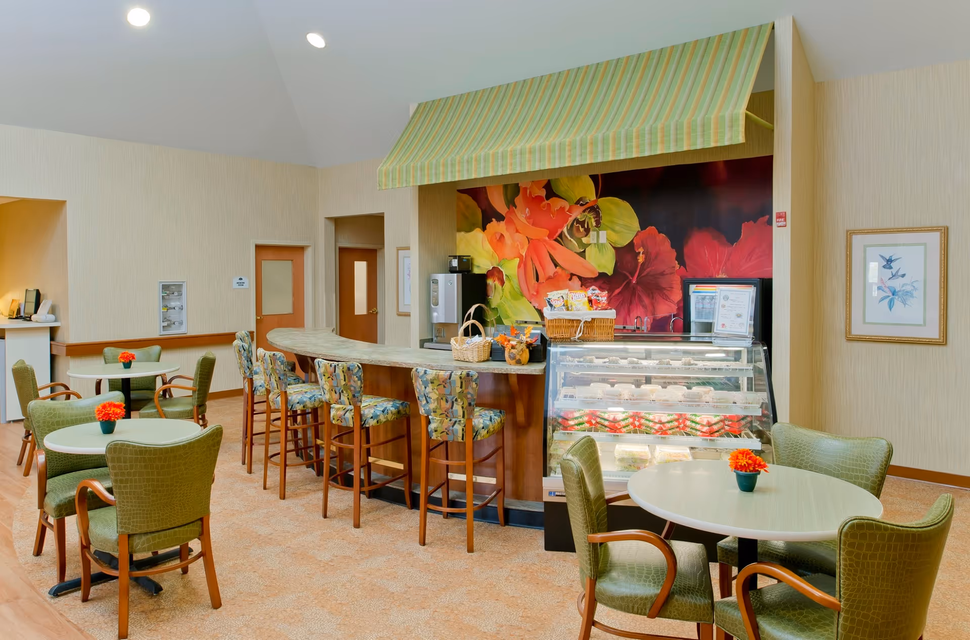 A cozy dining area in Lorien Mays Chapel featuring round tables with green cushioned chairs, a curved counter with floral-patterned bar stools, and a display case filled with desserts. The back wall has a large colorful floral mural and a striped green and yellow awning above the counter.