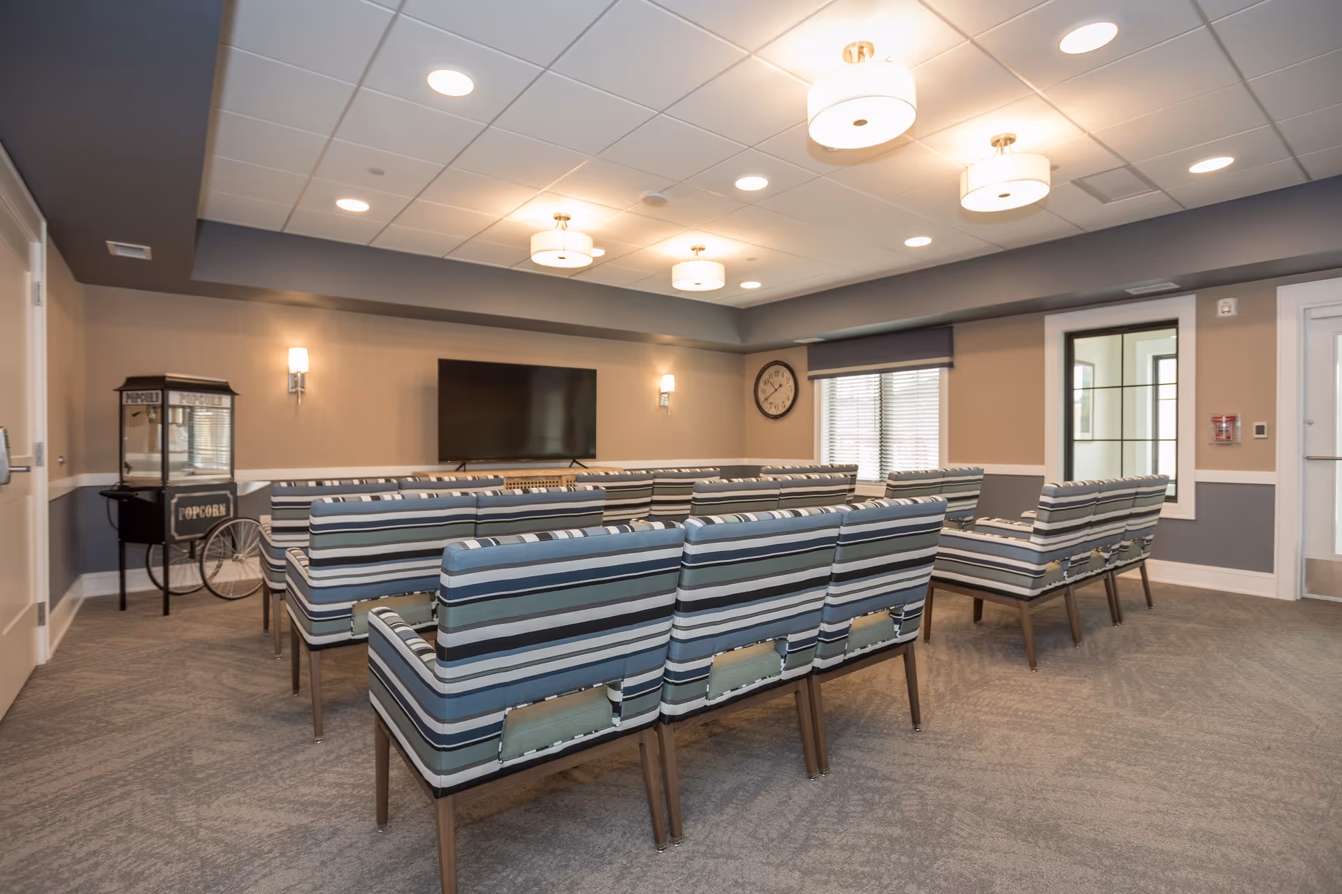 Communal media room with rows of striped upholstered chairs facing a wall-mounted TV and a popcorn cart.