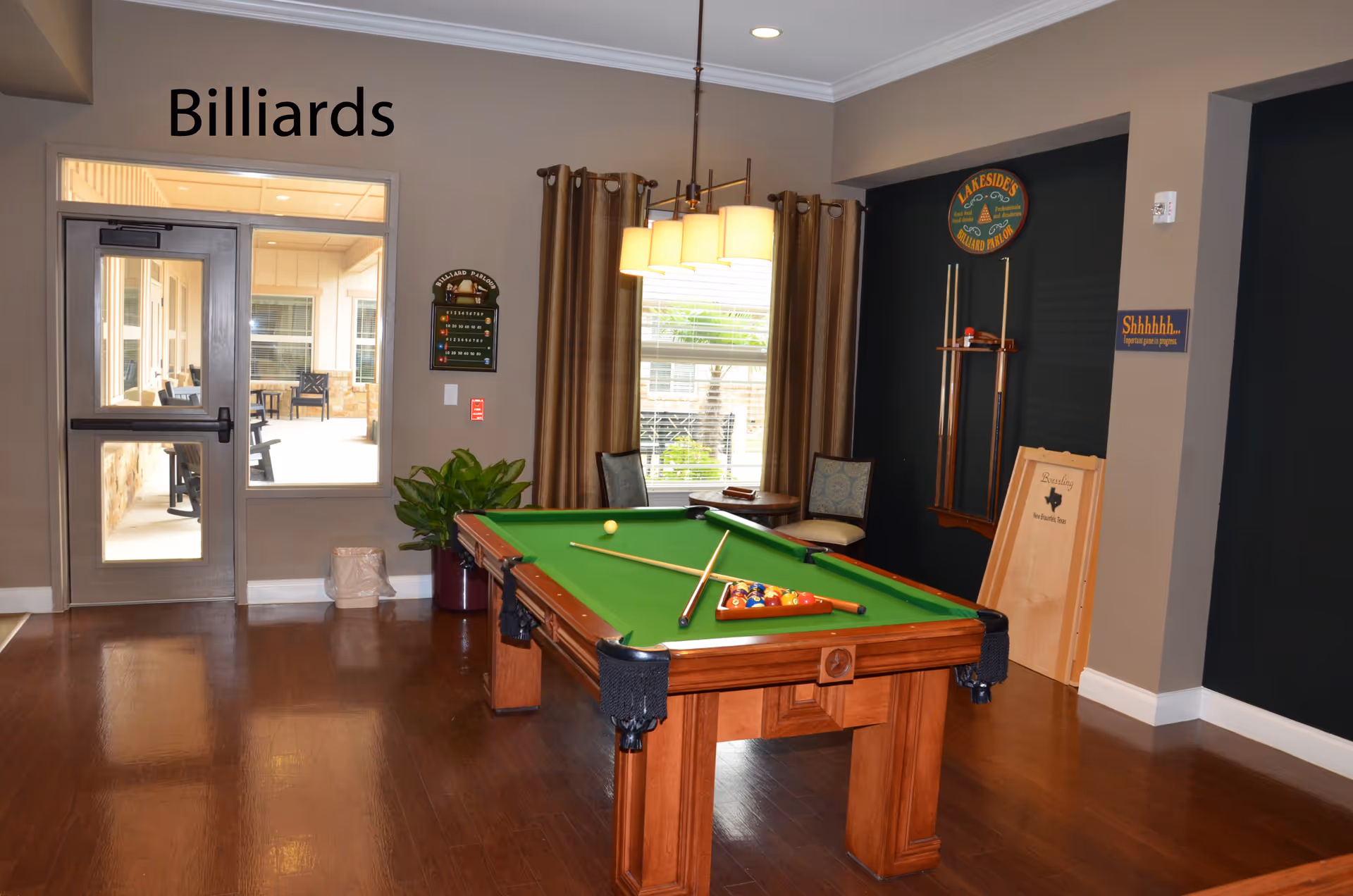 A billiards room with a green felt pool table in the center, two pool cues resting on the table, and a rack of billiard balls. The room has wooden flooring, beige walls, a window with brown curtains, and a door leading to an outdoor patio area. There are signs on the walls including one that says 'Billiards' and another that says 'Shhhhhh...'.
