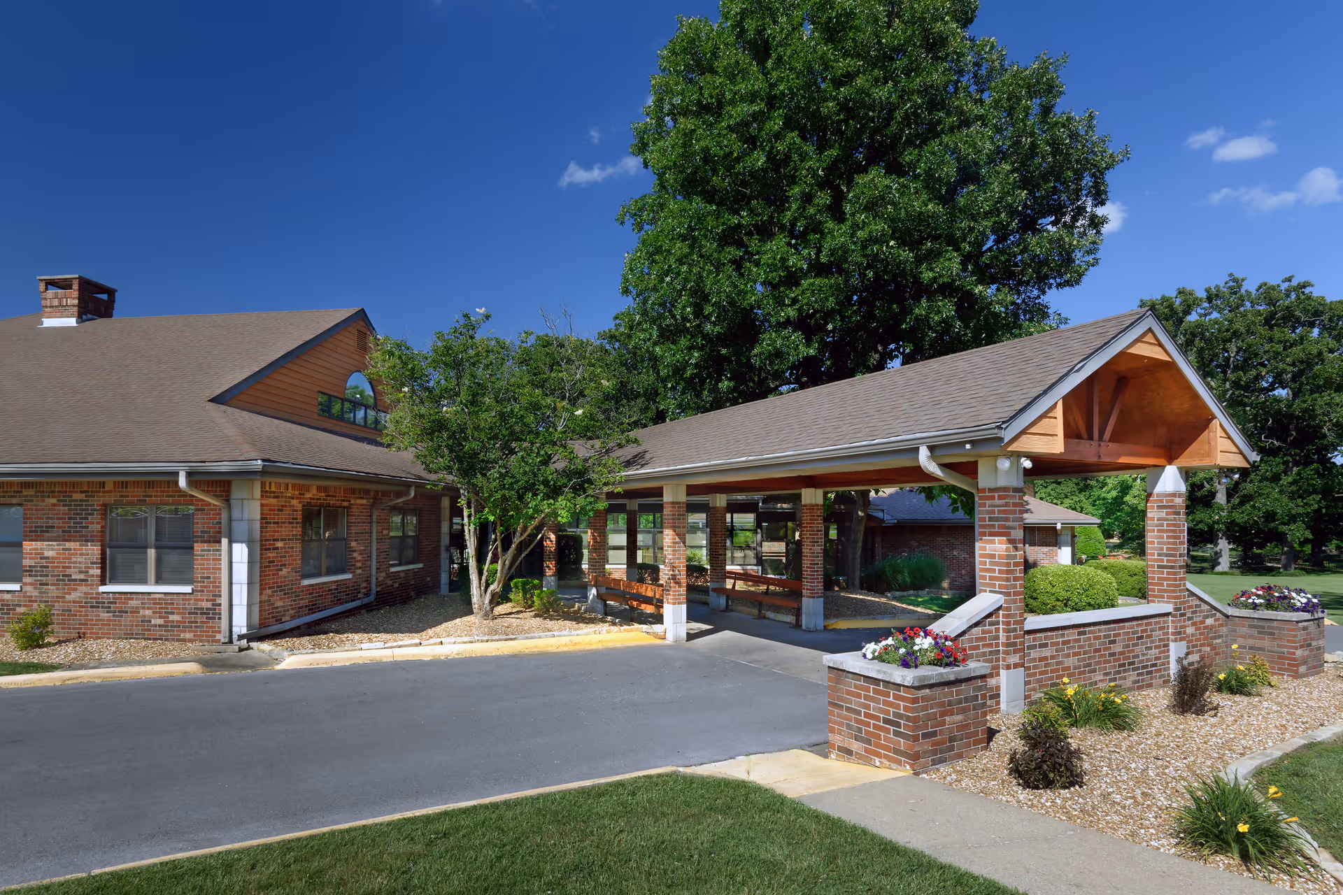 Covered brick porte-cochere and landscaped entrance in front of a residential care building under a clear blue sky.