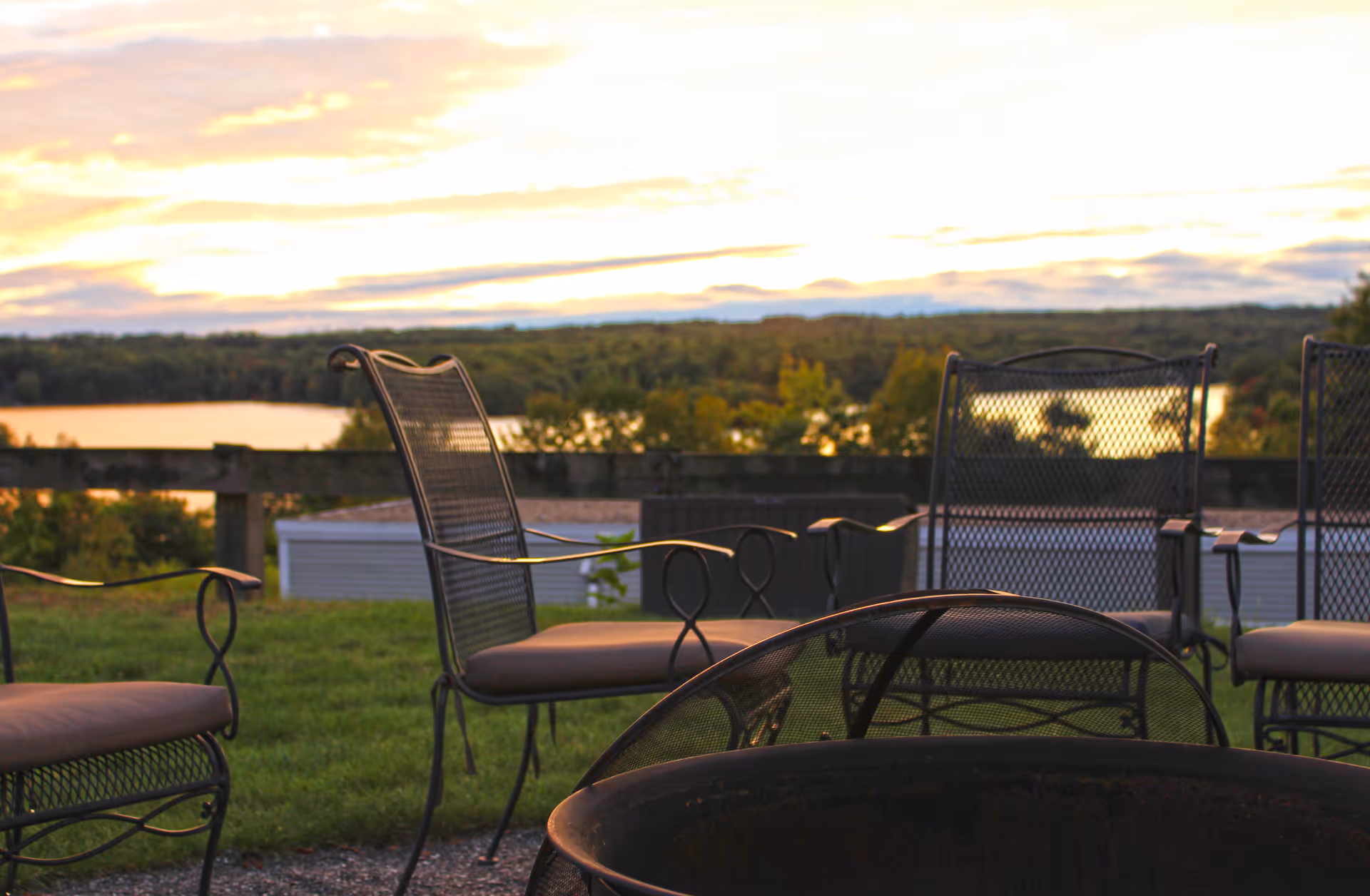 Outdoor seating area with metal chairs arranged around a fire pit, overlooking a scenic view of a lake and forested landscape at sunset.