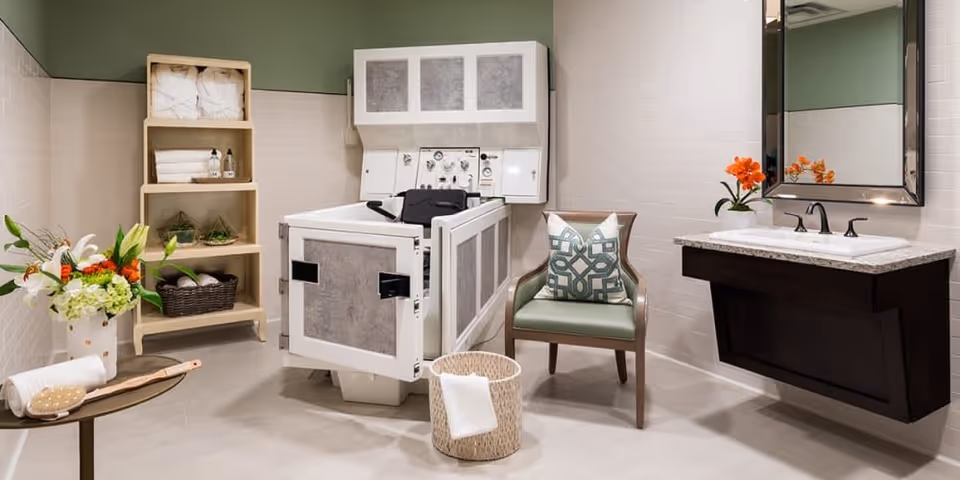 A clean and well-lit bathroom with a specialized bathing tub designed for assisted bathing, a wooden shelving unit with towels and toiletries, a green cushioned chair with a decorative pillow, a wall-mounted sink with a mirror above it, and floral arrangements adding a touch of color.