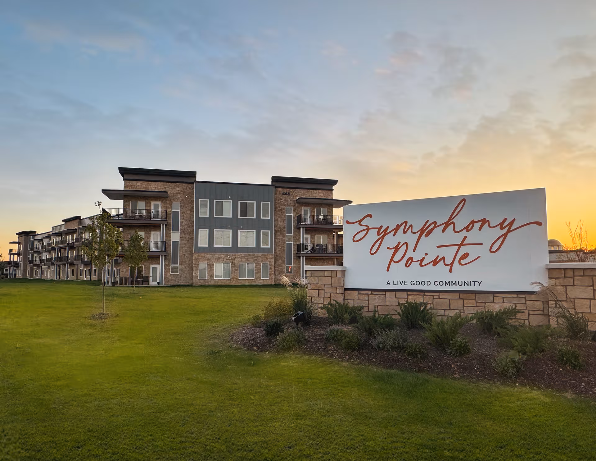 Exterior view of Symphony Pointe senior living community building at sunset with a large sign in front that reads 'Symphony Pointe A Live Good Community' surrounded by green grass and landscaping.