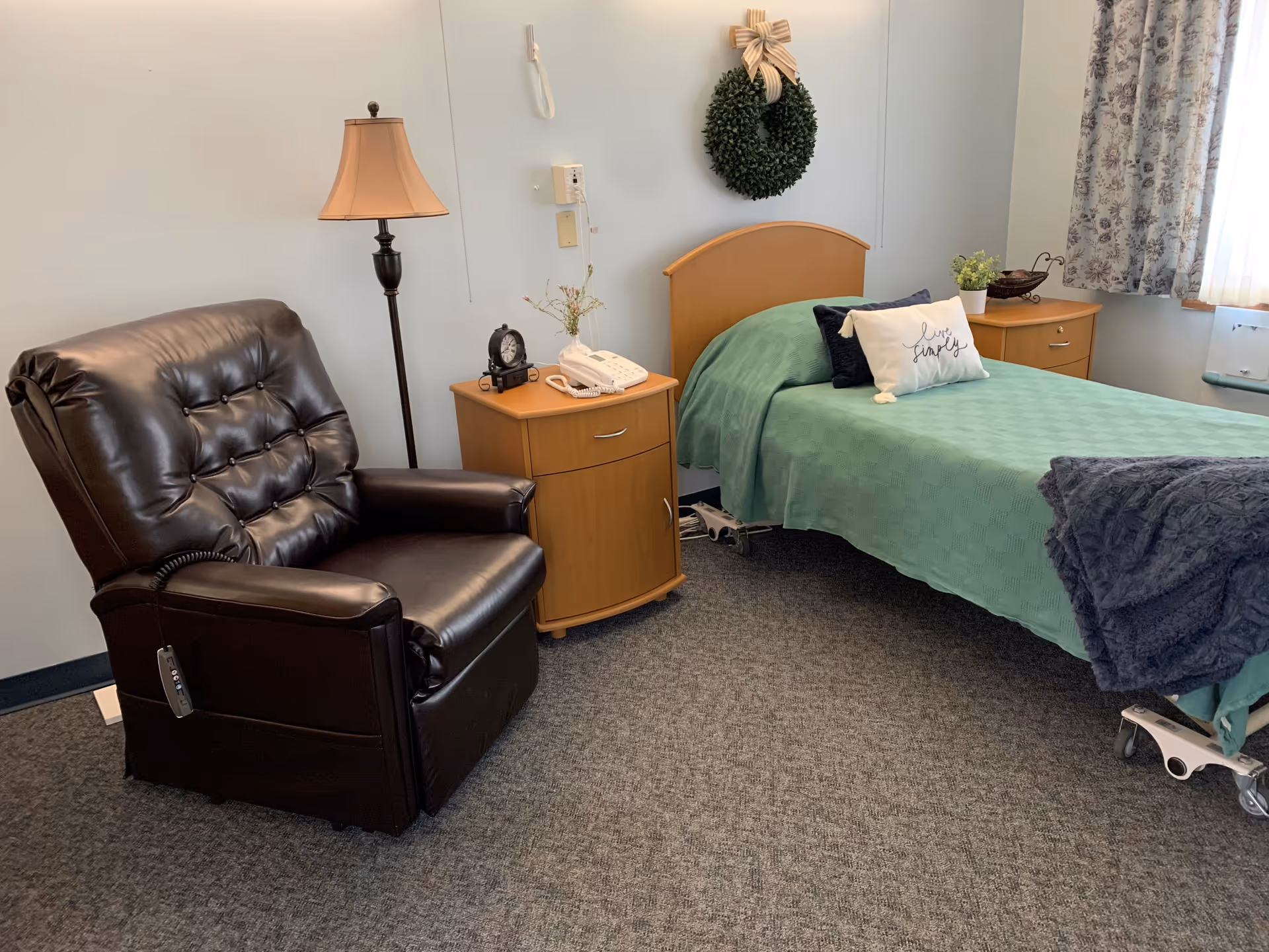 A cozy room with a single bed covered with a green blanket and decorative pillows, a dark brown leather recliner chair, a wooden nightstand with a lamp, telephone, and clock, a wreath hanging on the wall above the bed, and a window with floral curtains letting in natural light.