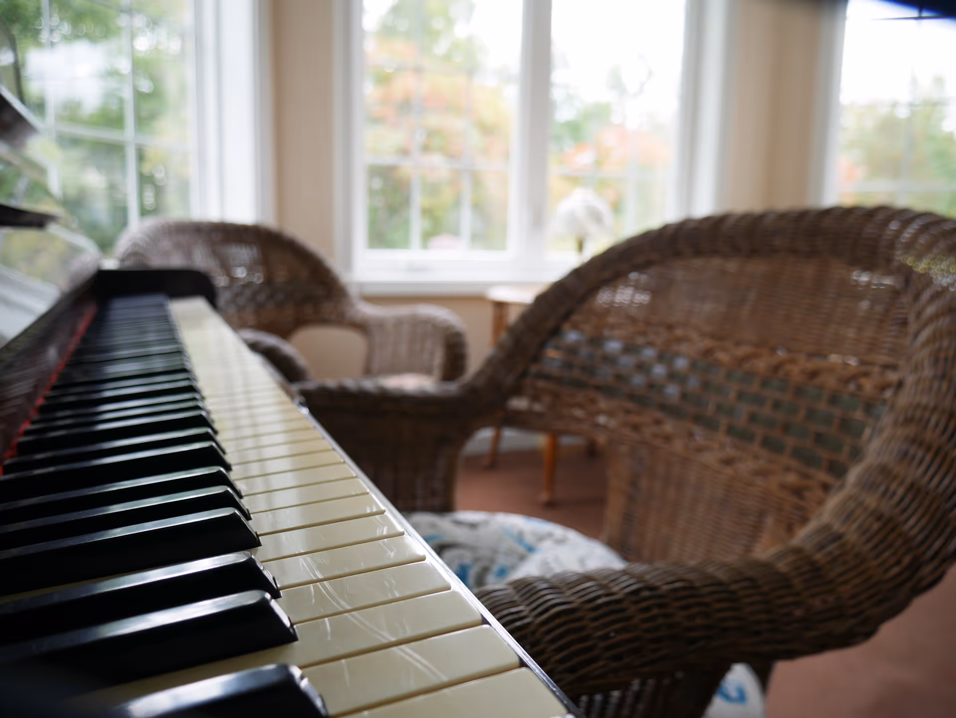 Close-up of piano keys in a sunlit room with wicker chairs and large windows in the background.
