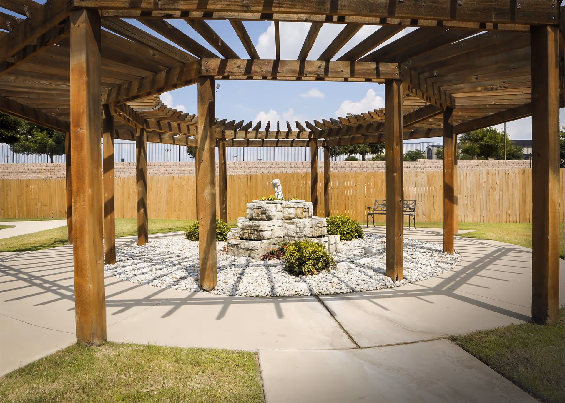 Outdoor seating area with a wooden pergola casting shadows on a circular stone and pebble garden bed with small bushes and a stone sculpture in the center, surrounded by a concrete pathway and grass, with a wooden fence and trees in the background under a partly cloudy sky.