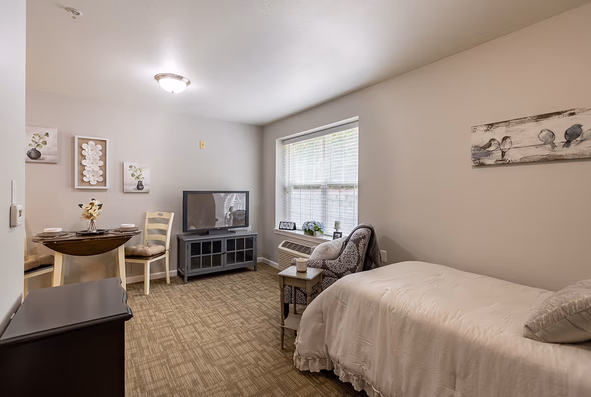 A cozy senior living room featuring a single bed with white bedding, a cushioned armchair with a side table, a flat-screen TV on a stand, a small round dining table with two chairs, and wall art including floral and bird-themed paintings. The room has a large window with blinds and beige walls and carpet.