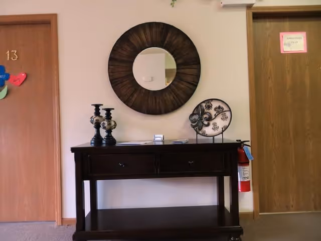 A hallway area with two wooden doors on either side, one labeled with the number 13 and decorated with colorful paper hearts. Between the doors is a dark wooden console table with two drawers, holding decorative items including two candlesticks and a decorative plate. Above the table is a round wooden-framed mirror. A fire extinguisher is mounted on the wall to the right of the table.