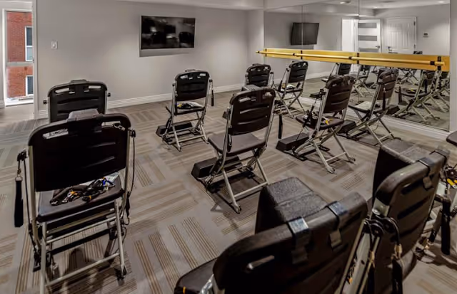 A fitness or exercise room with multiple black folding chairs arranged in rows facing a wall-mounted TV. The room has a mirrored wall with a ballet barre and a window letting in natural light. The floor is carpeted with a patterned design.