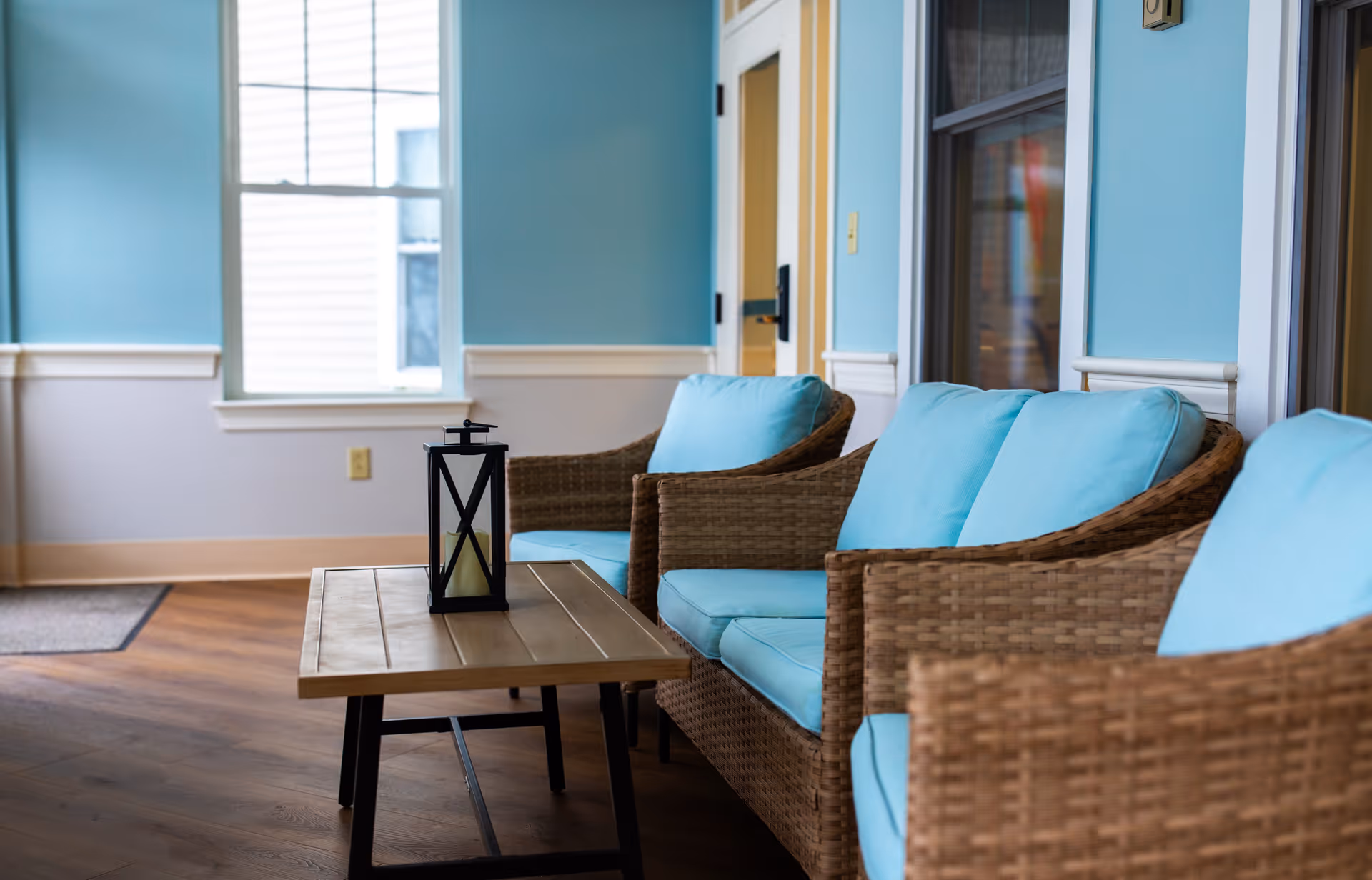 A cozy seating area with three wicker chairs featuring light blue cushions arranged around a wooden coffee table with a black lantern on top. The room has light blue walls, a window letting in natural light, and wooden flooring.