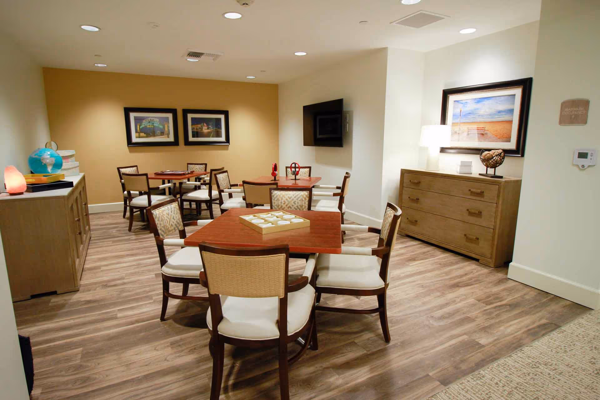 A well-lit common area with several wooden tables and cushioned chairs arranged for group activities or socializing. The room features wood flooring, a wall-mounted TV, framed artwork, a wooden dresser with decorative items, and a salt lamp on a side cabinet.