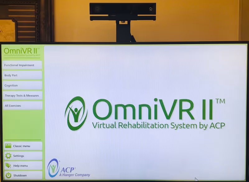 A computer screen displaying the OmniVR II Virtual Rehabilitation System by ACP interface, with menu options on the left side including Functional Impairment, Body Part, Cognition, Therapy Tests & Measures, and All Exercises. A camera or sensor device is mounted above the screen.