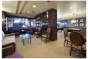 Cozy senior living community lounge with upholstered seating, round tables, a bookshelf and fireplace, and large windows.