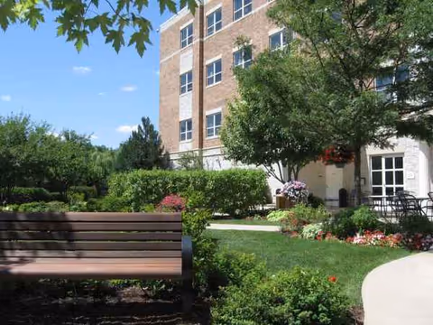 Outdoor garden area with a bench, lush landscaping, and a brick senior living building.