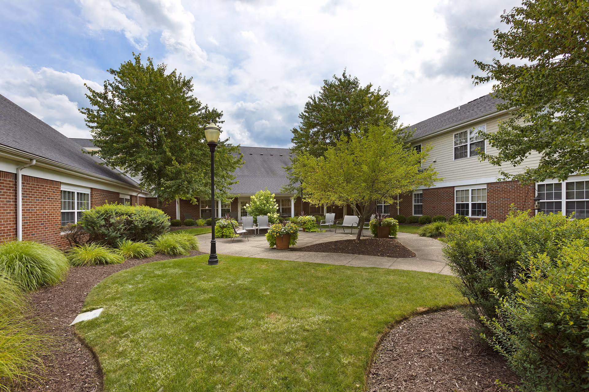 Outdoor courtyard area at Independence Village of Aurora featuring a well-maintained lawn, landscaped bushes, trees, a paved seating area with chairs and benches, and a lamppost under a partly cloudy sky.