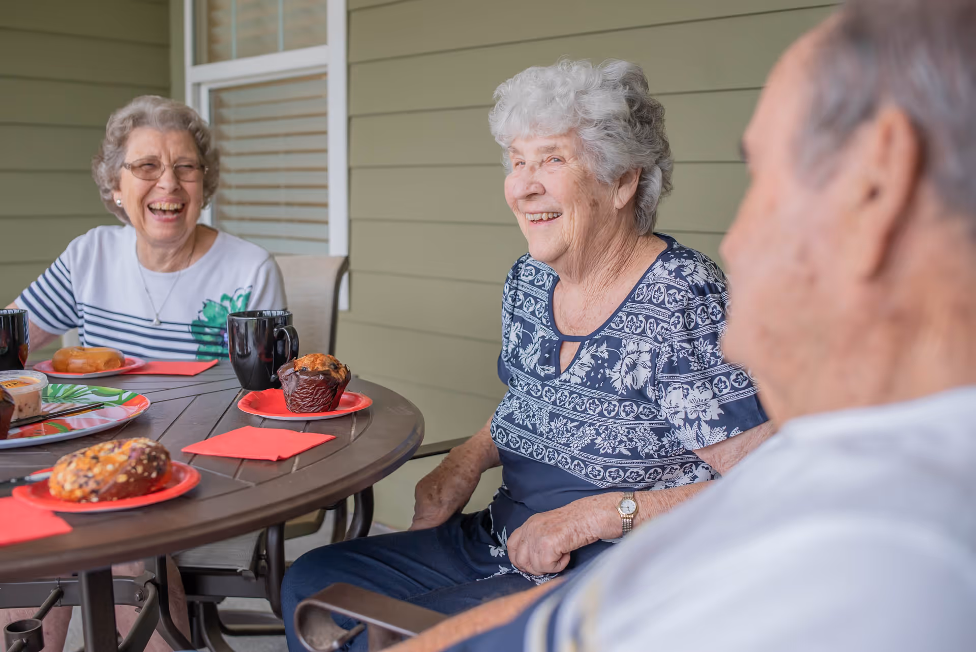 Three elderly people sitting around a round outdoor table with pastries and coffee cups, smiling and enjoying each other's company on a patio with green siding and a window in the background.