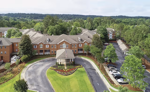 Aerial view of a multi-story brick senior living building with a covered front entrance, circular driveway, landscaped lawn, and adjacent parking surrounded by trees.